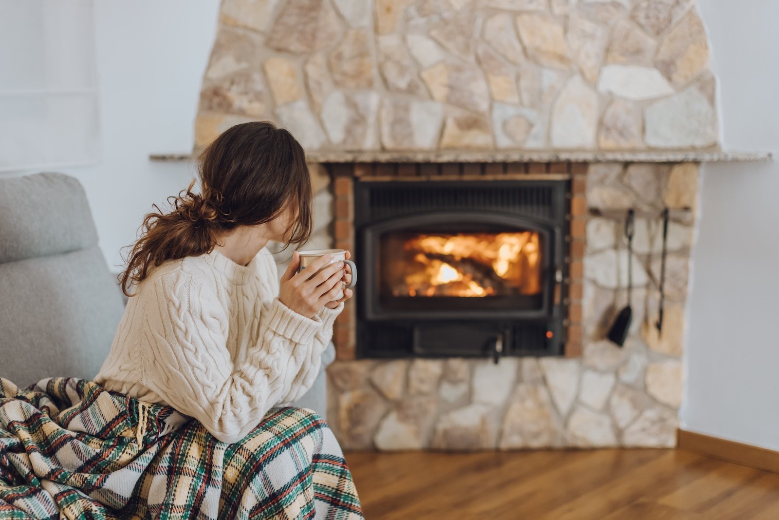 Young woman sitting at home by the fireplace with a hot tea or coffee mug and warming her hands, she is wearing white woollen sweater. Cold houses in Europe concept during energy and gas crisis.
